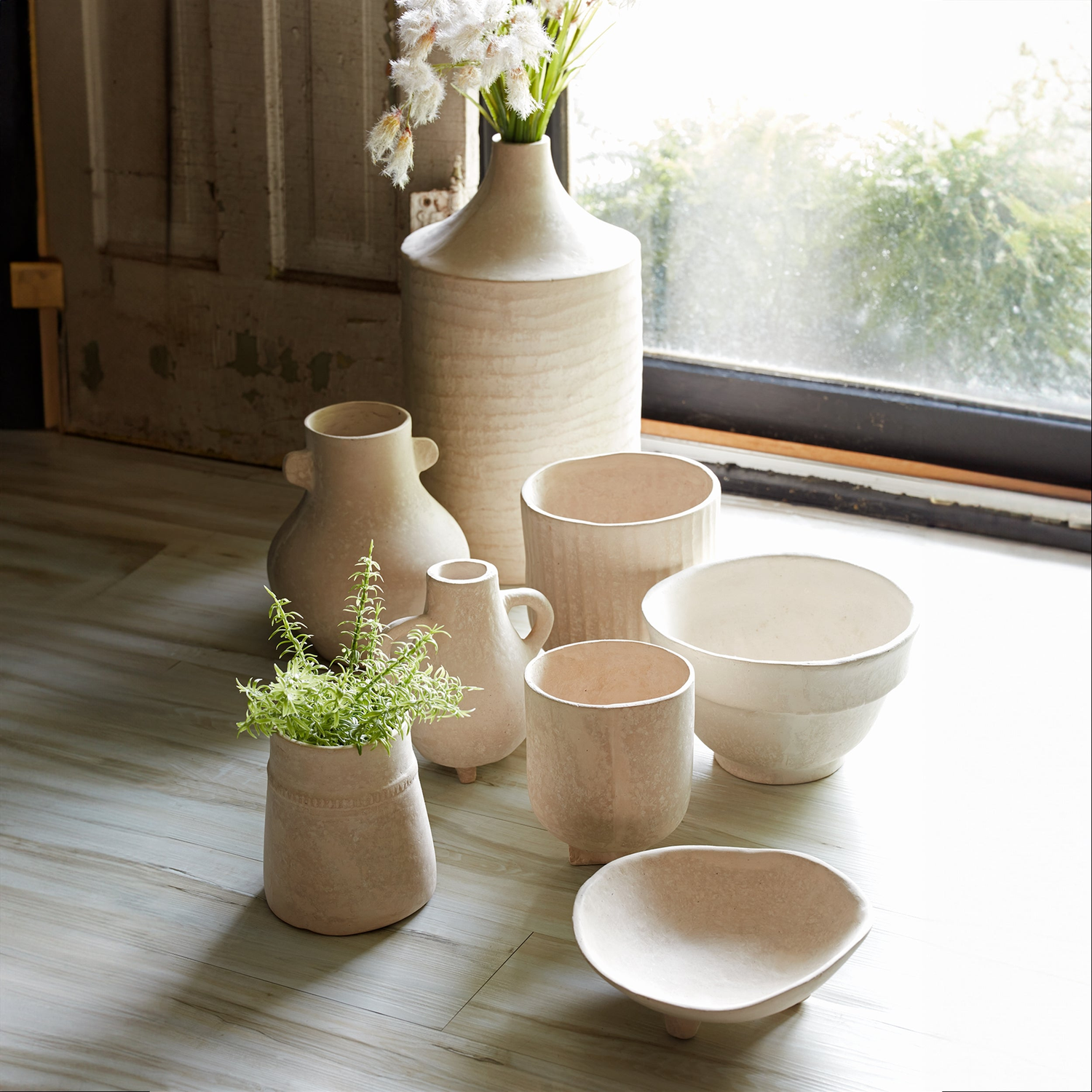 A collection of neutral-toned ceramic vases, bowls, and cups arranged on a wooden surface near a window. Some vases contain greenery and white flowers. The soft lighting and rustic wooden background create a cozy, natural aesthetic.